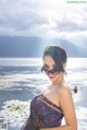 A woman in a bathing suit and sunglasses posing by the water.