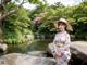 A woman in a kimono and hat standing by a pond.