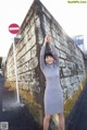 A woman in a gray dress leaning against a stone wall.