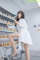 A woman in a white lab coat sitting on a stool in a pharmacy.